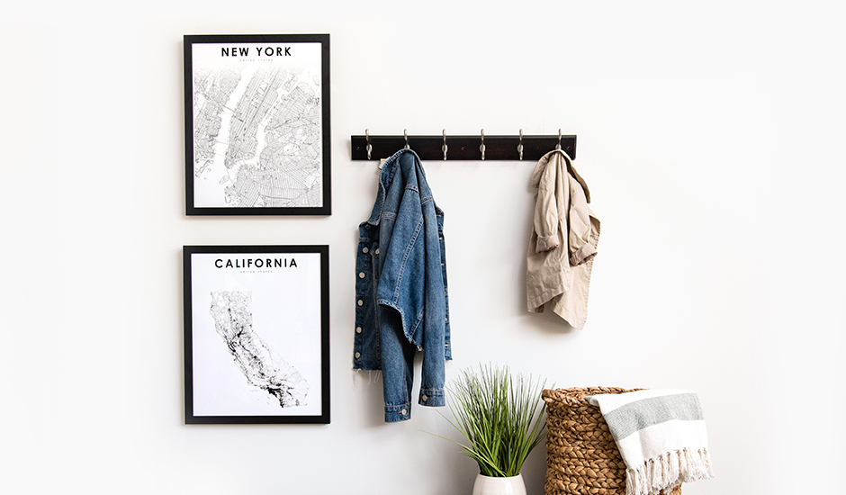 Two black framed photo prints of a map of California and New York hanging on a white wall next to a coat hanger with two jackets