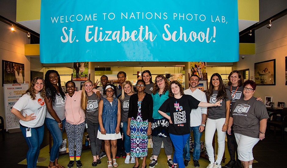 Nations Photo Lab employees gather in the lobby for a group photo standing under a banner for the St Elizabeth School